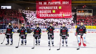 Eishockey-Spieler der Fischtown Pinguins stehen erschöpft und aufgereiht auf dem Eis, hinter ihnen zeigen die Fans eine 