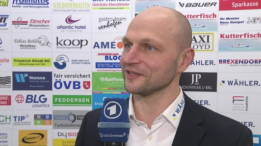 Eishockey-Manager Sebastian Furchner von den Fischtown Pinguins steht nach einem Spiel lächelnd vor einer Werbewand beim Interview.