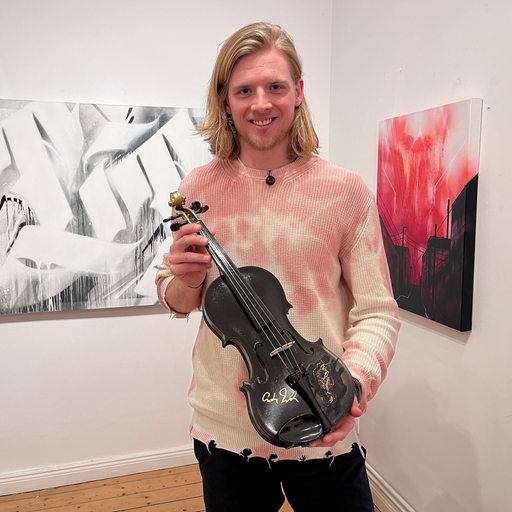 Pinguins-Spieler Moritz Wirth steht lächelnd in einer Kunstgalerie mit einer signierten schwarzen Geige in der Hand.