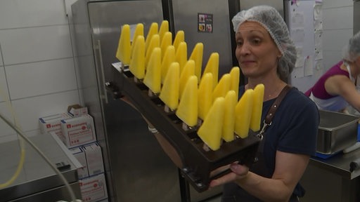 Eine Frau macht in einer Großküche Eis am Stiel und trägt ein großes Tablett mit gelben Eis vor sich her.