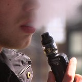 Ein Mann hält ein Vape in der Hand und pustet aus dem Mund Dampf aus.