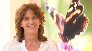 Frau mittleren Alters mit langen Locken strahlt vor übergroßer Schmetterlingsnachbildung in die Kamera