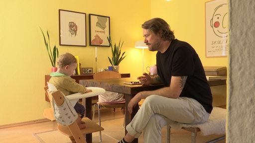 Es ist ein Mann zu sehen, der gegenüber von einem Kleinkind in einem Wohnzimmer sitzt.