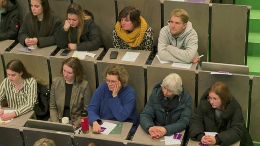 Personen verschiedenen Alters sitzen im Hörsaal und schauen nach vorn, sie haben Flyer und Handys vor sich auf den Tischen
