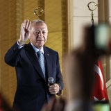 Ein Mann steht mit einem Mikrofon in der Hand. Er hält die andere Hand nach oben wie zum Gruße. Im Hintergrund Flaggen der Türkei.