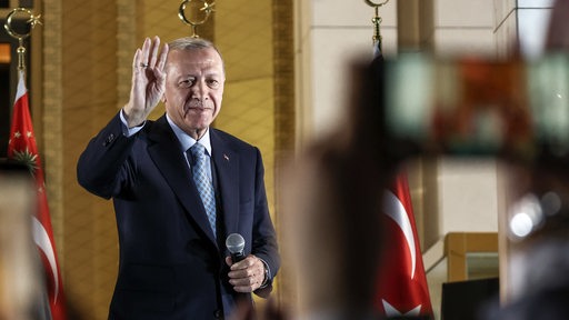 Ein Mann steht mit einem Mikrofon in der Hand. Er hält die andere Hand nach oben wie zum Gruße. Im Hintergrund Flaggen der Türkei.