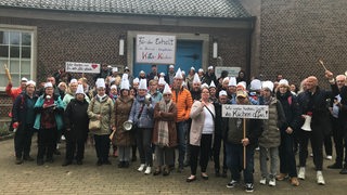 Eine Personengruppe protestiert für den Erhalt der Kita-Küchen, einige haben Plakate, Kochlöffel oder Kochmützen dabei