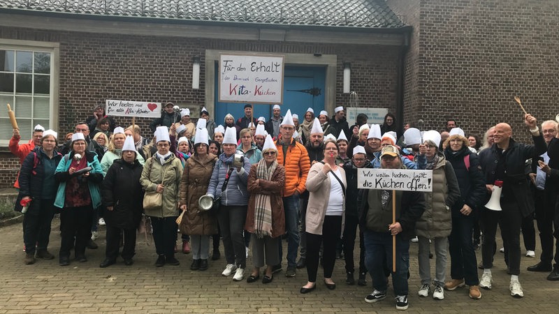 Eine Personengruppe protestiert für den Erhalt der Kita-Küchen, einige haben Plakate, Kochlöffel oder Kochmützen dabei