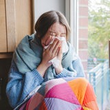 Eine kranke junge Frau sitzt am Fenster und schnaubt sich die Nase.