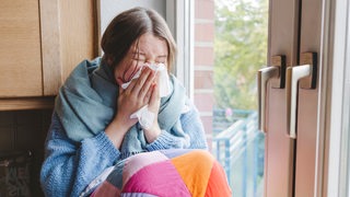 Eine kranke junge Frau sitzt am Fenster und schnaubt sich die Nase.