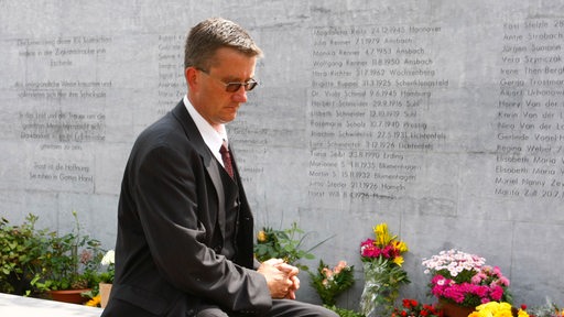 Ein Mann im Alter von 40 bis 45 Jahren etwa sitzt auf einem Steint vor einer Gedenktafel. Neben dem Mann ein Gehstock. Auf der Tafel stehen viele Namen. Davor stehen Blumen. 