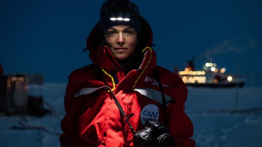 Eine Frau steht in einer roten Jacke mit Kopflampe und Kamera im Schnee.