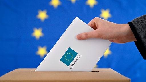 Ein Wahlbrief wird in eine Wahlurne gesteckt, im Hintergrund eine EU-Flagge. 
