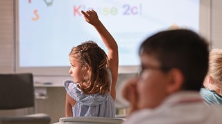 Ein Mädchen in einer Grundschule meldet sich, im Hintergrund ein White-Board.