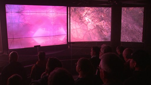 Zuschauer sitzen in einem dunklen Raum und blicken auf große Bildschirme in der Extremwetterausstellung im Klimahaus 
