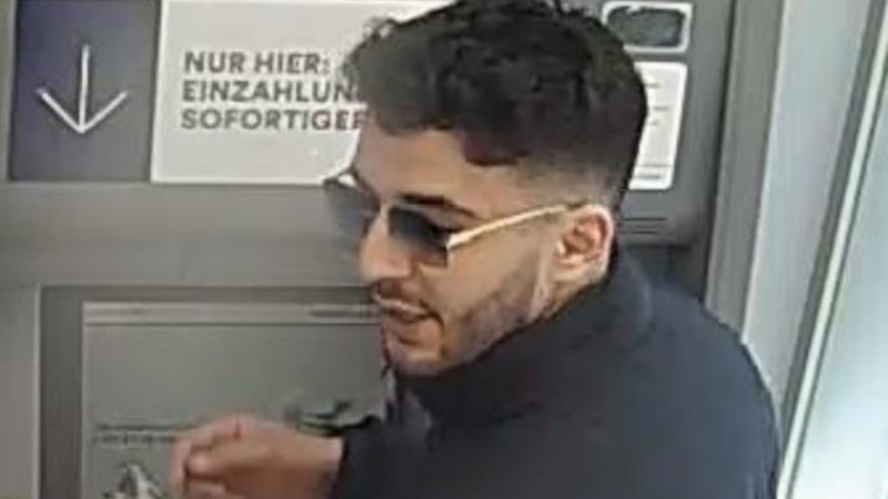 Ein dunkelhaariger Mann mit Drei-Tage-Bart und Sonnenbrille steht vor einem Geldautomat