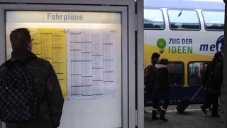 Ein Mann steht im Bahnhof an dem Fahrplanaushang. Im Hintergrund steht ein Metronom.