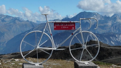 Ein weißes Fahrrad mit einem Schild auf dem "COL DE LA LOZE" steht. Im Hintergrund sind Berge zu erkennen.