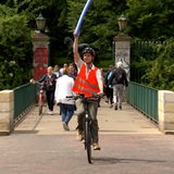 Ein junger Mann mit einer Warnweste und einem Fahrradhelm fährt auf seinem Rad über eine Brücke. In der Hand hält er eine Pool-Nudel und reckt sie in die Luft.