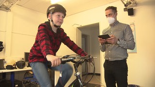 Ein Junge sitzt mit Helm auf ein Fahrrad-Simulator, neben ihm ein Forscher mit Tablet.