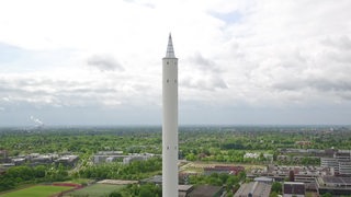 Der Bremer Fallturm sieht aus wie ein überdimensionaler Bleistift, der in den Himmel ragt. Im Hintergrund ist das Uni-Gelände zu sehen.