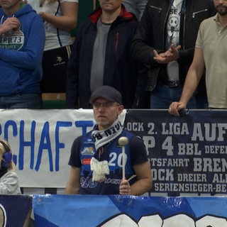 Ein Mann mit Trikot und Schal der Eisbären Bremerhaven steht vor einem Banner und haut mit einem Schlegel auf eine Trommel
