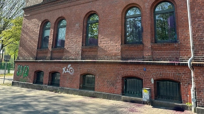 Eine rote Backsteinwand eines Hauses, die Fenster und der Boden davor sind mit lila Farbe beschmiert.