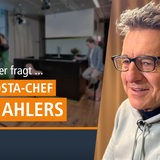 Gauhaariger Mann mit dunkler Brille und Kaputzen-Shirt vor Fernsehstudio-Kulisse