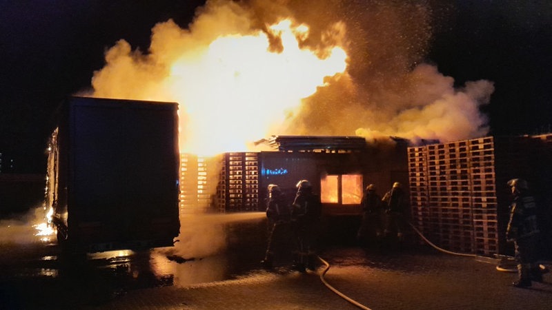 Feuerwehrleute stehen mit einem Schlauch vor brennenden Paletten.