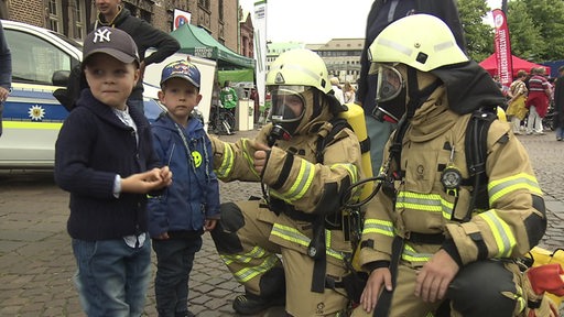 Zwei Feuerwehrleute in Arbeitskleidung sprechen bei einem Aktionstag mit Kindern.