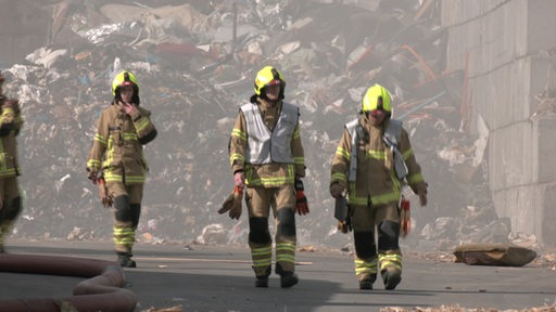 Vier Feuerwehr-Leute laufen vor einem Brennenden Müllberg.