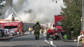 Ein Feuerwehrmann läuft auf einen Brand zu. Auf einer Recyclinganlage brennt Sperrmüll, der bereits mit einer Drehleiter gelöscht wird.