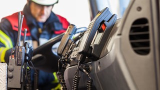 Funkgeräte sind in einem Feuerwehrfahrzeug zu sehen, im Hintergrund ein Feuerwehrmann.