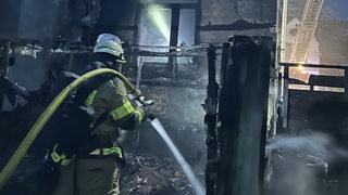 Ein Feuerwehrmann steht vor einem ausgebrannten Haus und spritzt Wasser aus dem Löschschlauch.