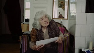 Die Holokaustüberlebene Lore Kübler sitzt in einem Sessel in ihrer Wohung und hält Papiere in der Hand.