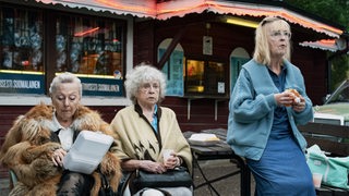 Filmszene aus "Ladies of Steel", 3 ältere Frauen sitzen mit Fastfood auf einer Bank in der Stadt