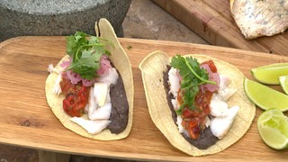 Ein Holzbrett auf dem zwei Fisch-Tacos liegen mit Gemüse und frischer Petersilie als Toppings und Limettenspalten daneben.