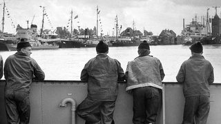 Historisches Schwarzweißbild. Vier Fischer blicken auf Boote im Fischereihafen Bremerhaven.
