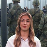 Eine junge Frau mit langen dunklen Haaren vor einem Bild einer Militärübung.
