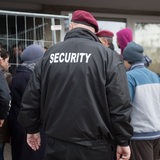 Rückenansicht eines Security-Mitarbeiters inmitten von Flüchtlingen vor einem Flüchtlingsheim