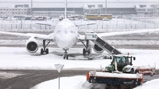 Ein Schneeräumer fährt im Schneetreiben auf dem Flughafen vor einer Maschine entlang.