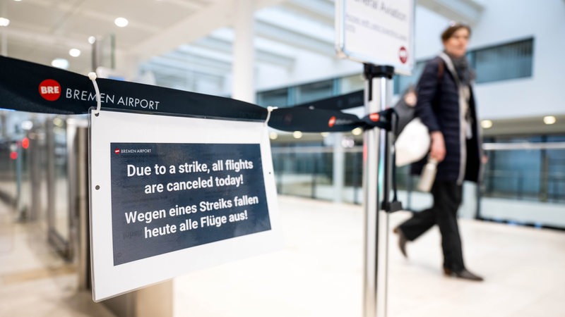 Ein Schild informiert über den Streik am Flughafen Bremen. Im Hintergrund läuft eine Frau mit einer Umhängetasche an dem Schild vorbei.