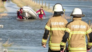 Zwei Feuerwehrmänner stehen vor einer überfluteten Salzwiese. Im Hintergrund sind Einsatzkräfte mit einem Rettungsboot zu erkennen.
