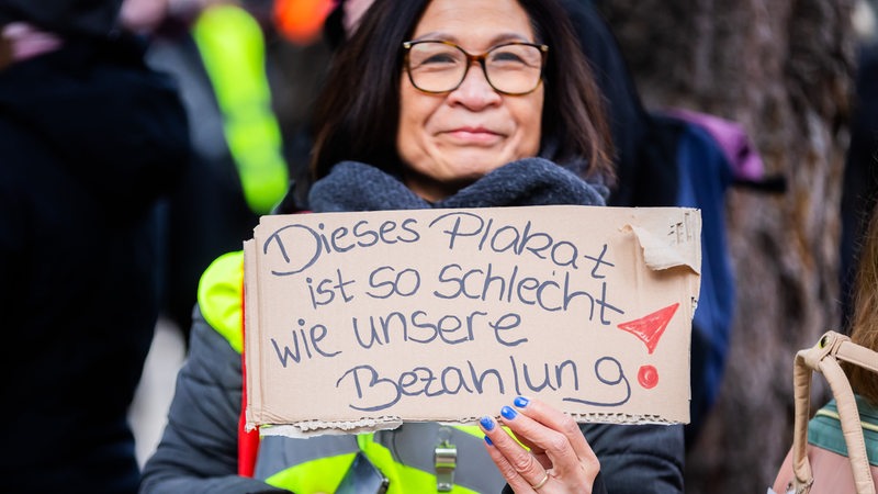 Eine Frau mit Warnweste und Brille zeigt ein Pappschild mit der Aufschrift "Dieses Plakat ist so schlecht wie unsere Bezahlung!"