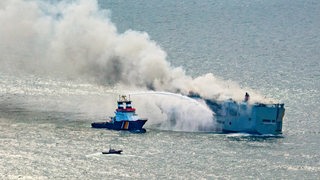 Ein Schiff richtet eine Wasserkanone auf einen brennenden Frachter. 