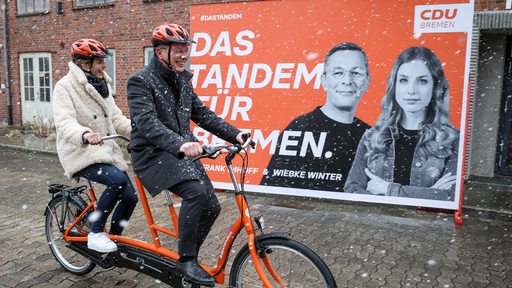 Ein Mann und eine Frau fahren auf einem Tandem mit Schutzhelmen auf dem Kopf. Im Hintergrund ein Wahlplakat mit Frank Imhoff und Wiebke Winter. 