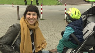 Eine junge Frau Frau lacht in Richtung Kamera und kniet neben einem Kinderbuggy. In dem Buggy sitzt ein kleines Kind mit einem buntem Fahrradhelm.
