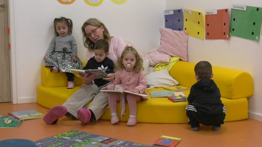 Eine Frau sitzt mit Kindern auf einer kleinen Couch und liest ein Buch.