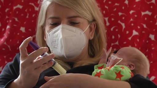 Eine junge Frau mit FFP2-Maske gibt einem Neugeborenem Nahrung durch eine Sonde. 