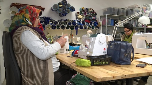Zwei Frauen sitzen an einem Tisch in einer Nähwerkstatt. Auf dem Tisch stehen Nähmaschinen.